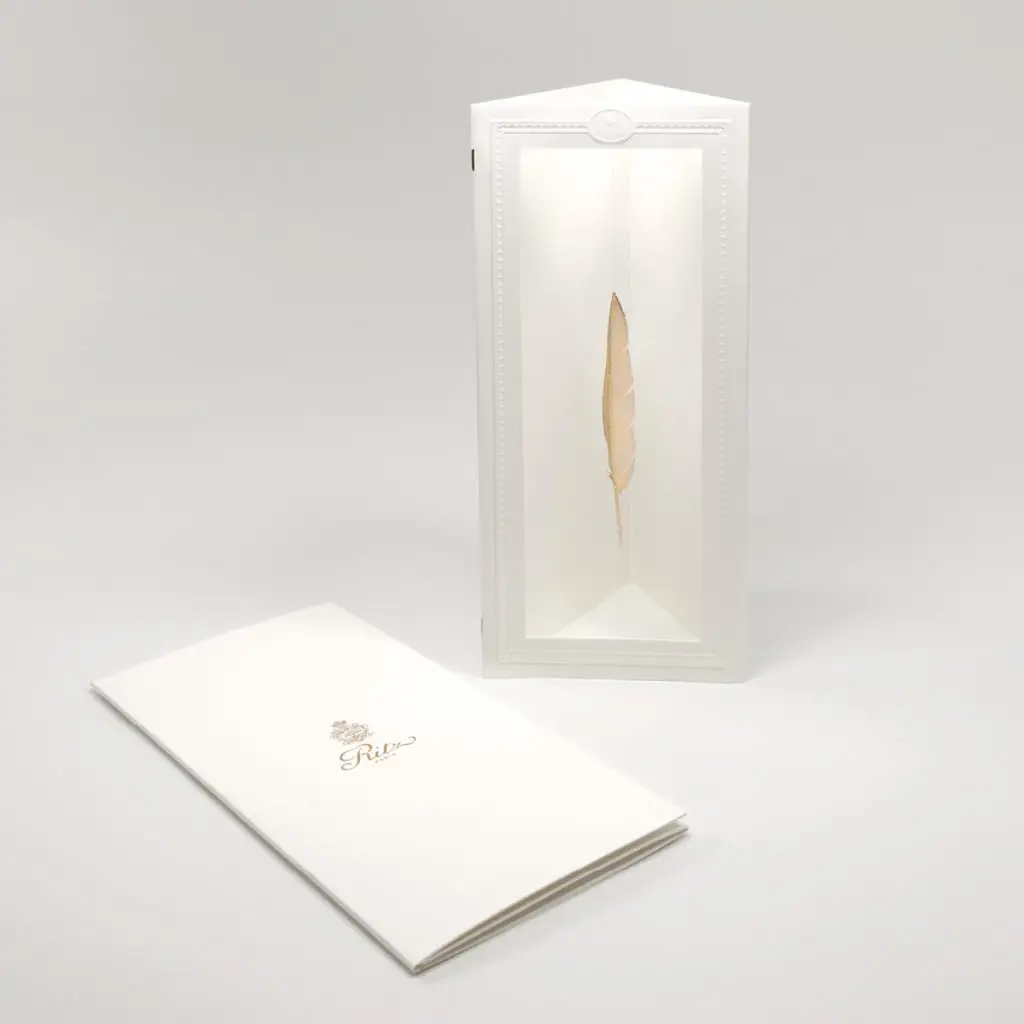Carte de vœux du Ritz Paris : écrin triangulaire en papier blanc gaufré présentant une plume en métal doré, à côté de la carte pliée marquée du logo doré