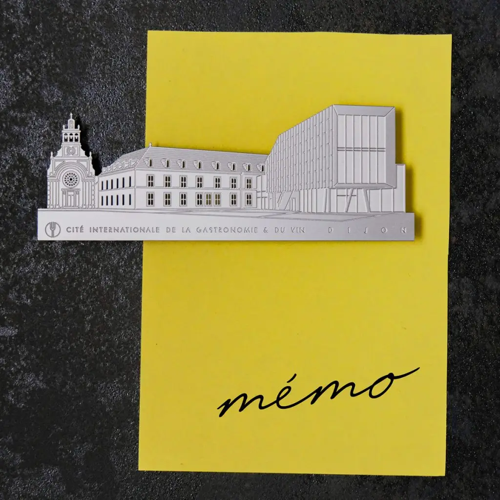 Magnet en métal argenté représentant le bâtiment de la Cité internationale de la gastronomie et du vin, posé sur un carnet jaune