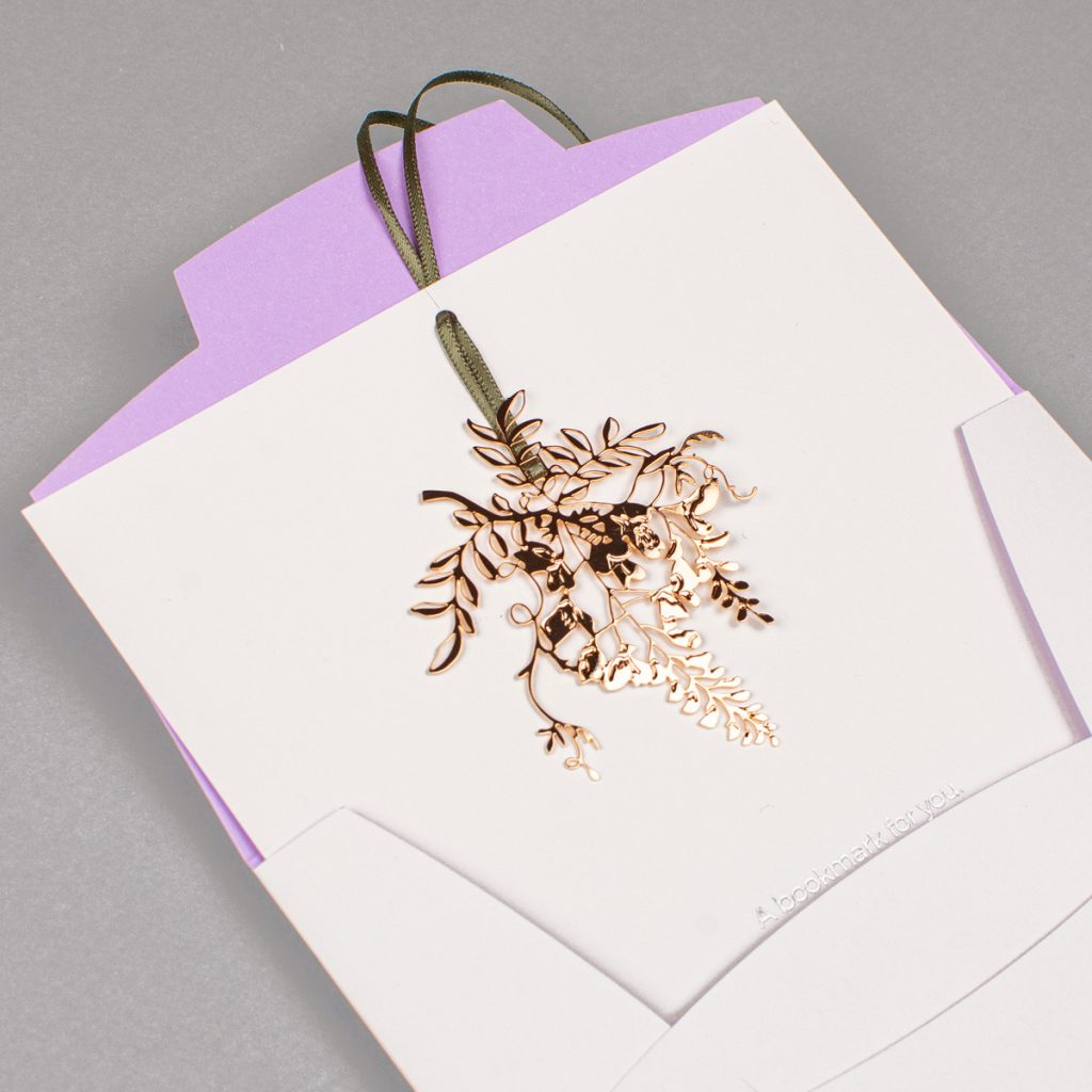 Marque-page en métal or rose ajouré en forme de glycine, glissé dans une pochette ivoire à l'intérieur lilas